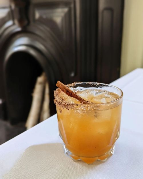 A glass of cocktail with ice, garnished with a cinnamon stick and a sugared rim, set against a dark background.