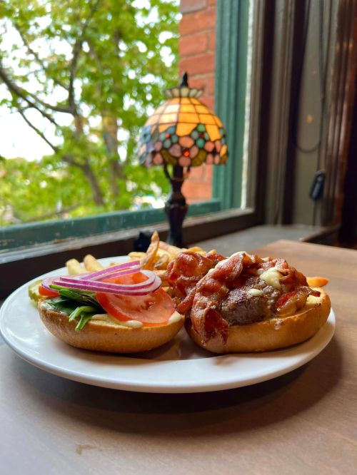 A plate with a burger topped with bacon, lettuce, tomato, and onion, alongside fries, near a window with a lamp.