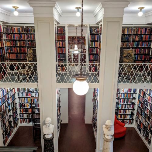 A spacious library with tall bookshelves, a central chandelier, and a red chair, featuring classical sculptures.