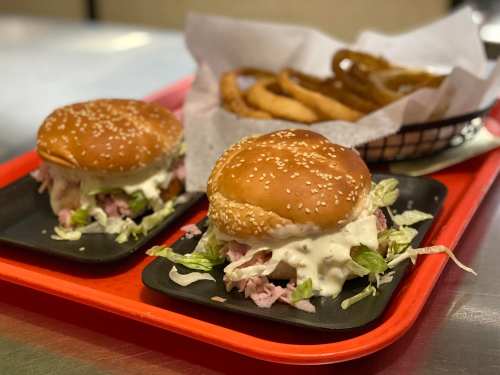 Two sesame seed burgers with lettuce and sauce on black plates, served with a side of crispy onion rings on a red tray.