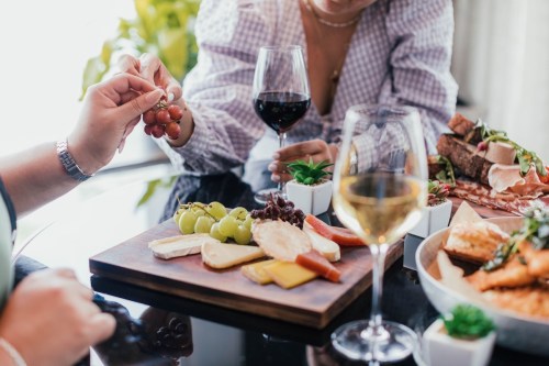 A charcuterie board with cheese, grapes, and wine glasses, as two people enjoy a meal together.