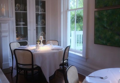 A cozy dining room with a round table set for four, featuring white tablecloths and a window with natural light.