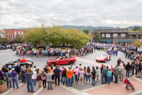 A crowd gathers around a parade with cars and performers in a small town square, surrounded by trees and buildings.