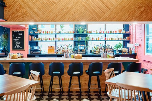 A modern bar with a wooden counter, high stools, and shelves filled with colorful bottles in a bright, stylish setting.