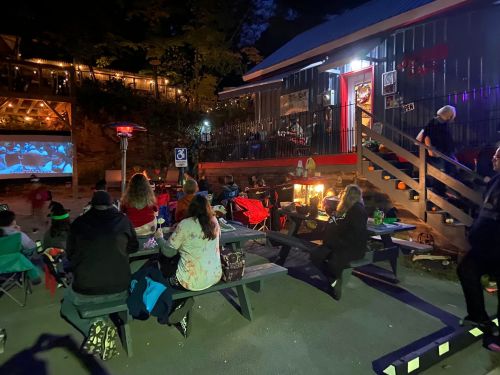 A group of people sitting around a fire pit at night, watching a movie on a screen outside a rustic building.