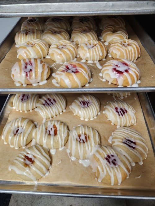 Freshly baked pastries drizzled with icing and filled with fruit jam, arranged on baking trays.