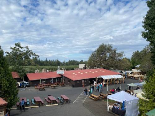 A view of a rustic outdoor market with red-roofed buildings, picnic tables, and people enjoying the space under a cloudy sky.