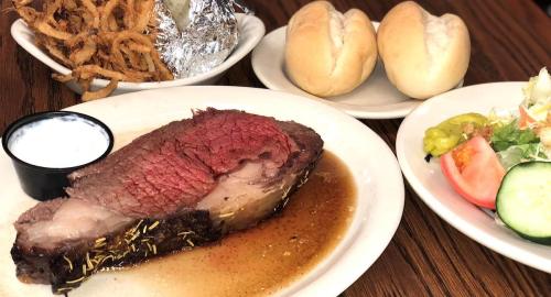 A plate with sliced roast beef in gravy, a side salad, two rolls, and crispy onion strings.