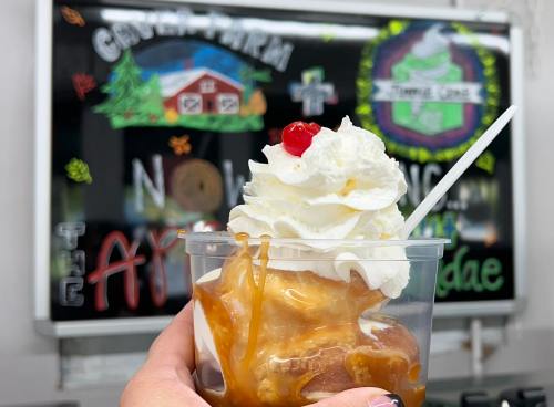 A hand holding a cup of ice cream topped with whipped cream, a cherry, and caramel sauce, with a colorful sign in the background.