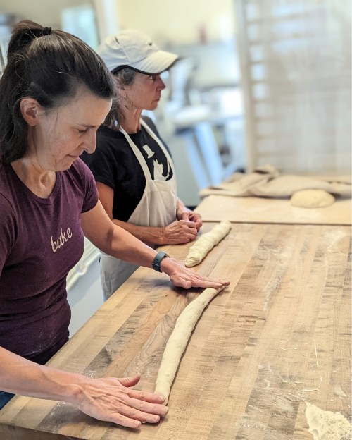 Two women are shaping dough on a wooden table in a bright kitchen. One is rolling the dough into a long shape.