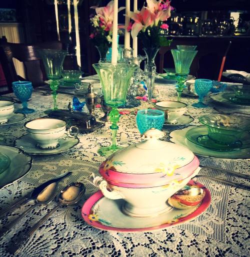 A beautifully set dining table with vintage china, glassware, and floral decorations on a lace tablecloth.
