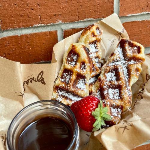 Two golden-brown waffles dusted with powdered sugar, served with a strawberry and a small jar of chocolate sauce.