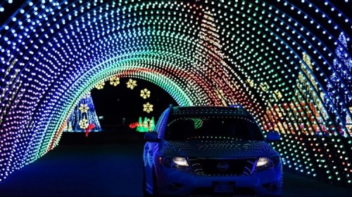 A car drives through a colorful light tunnel decorated for the holidays, with festive patterns and shapes.