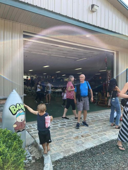 A busy market entrance with people walking, a child holding a drink, and a cartoon character sign nearby.