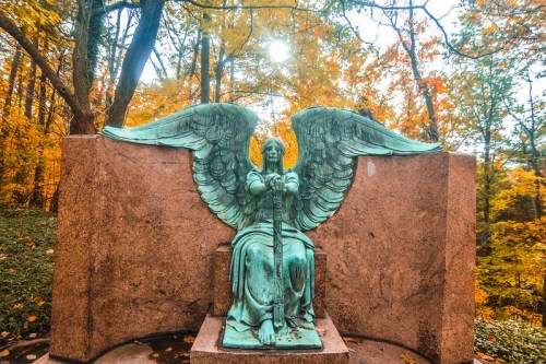 A green angel statue with wings sits on a stone pedestal, surrounded by vibrant autumn foliage and soft sunlight.