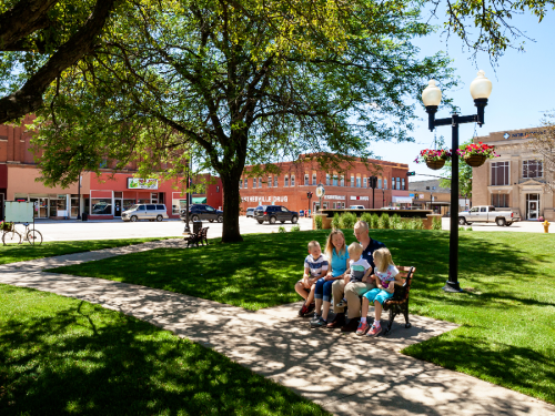 A family sits on a bench in a park, surrounded by trees and buildings, enjoying a sunny day.