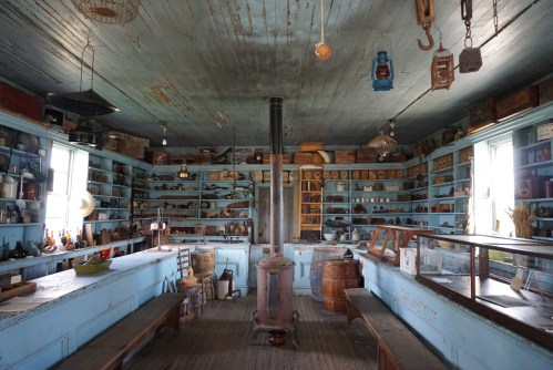 A vintage store interior with blue walls, wooden shelves filled with jars and tools, and a central wood stove.