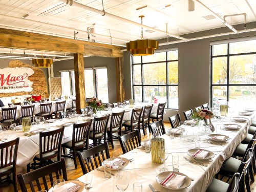 A spacious dining room with long tables set for a meal, large windows, and modern decor featuring wooden beams.
