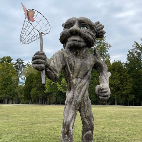 A wooden sculpture of a whimsical figure holding a butterfly net, set in a grassy area with trees in the background.