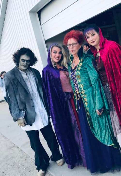 A group of four people in costumes, including a zombie and three women in colorful cloaks, posing together outdoors.