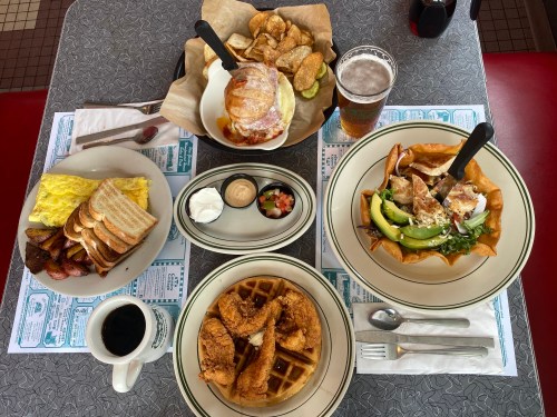 A table spread with breakfast and lunch items: eggs, toast, sausage, chicken, waffles, salad, and a drink.
