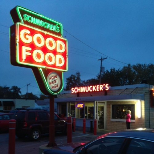 Neon sign for Schmucker's Good Food at dusk, featuring a diner with a customer at the window and parked cars nearby.