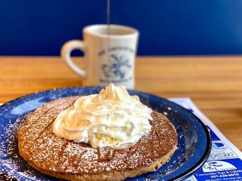 A large pancake topped with whipped cream and powdered sugar, with a coffee mug in the background.