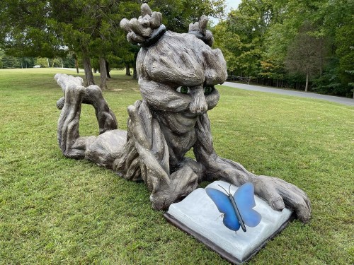 A whimsical tree-like sculpture of a creature reading a book with a blue butterfly on the pages, set in a grassy park.