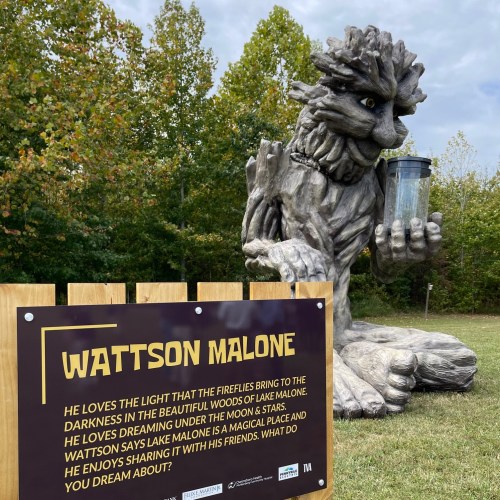 A whimsical tree sculpture named Wattson Malone, holding a lantern, surrounded by greenery and a sign with his name.