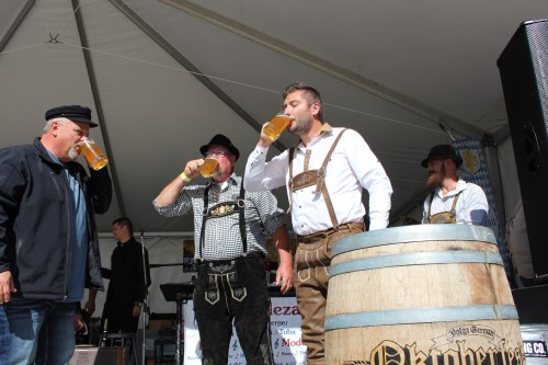 Four men in traditional attire enjoy beer at an Oktoberfest event under a tent, with a wooden barrel in the foreground.