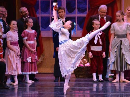 A young ballerina performs a graceful leap in a festive ballet scene, surrounded by other dancers in elegant costumes.
