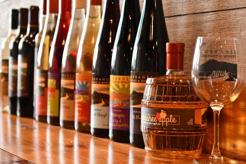 A row of various wine bottles and a glass on a wooden shelf, showcasing a selection from a vineyard.