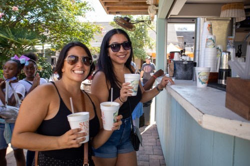 Two women in sunglasses smile while holding drinks at a vibrant outdoor café, with people and greenery in the background.