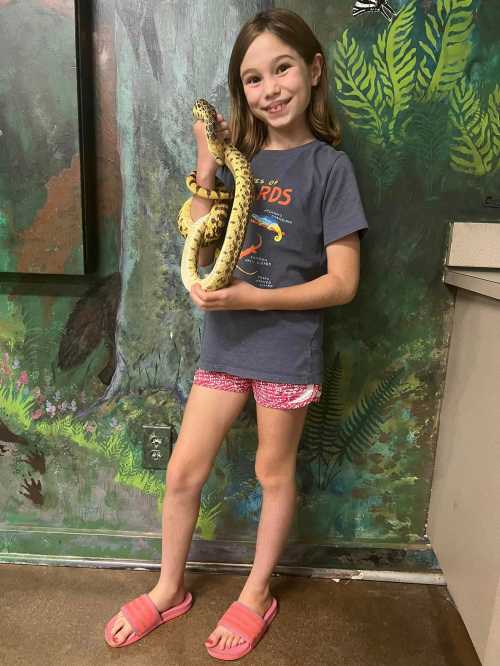 A smiling girl holds a snake while standing in front of a colorful, nature-themed mural. She wears a t-shirt and shorts.