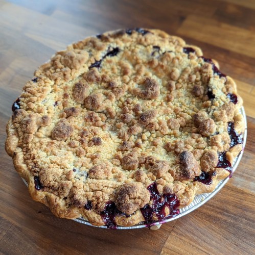 A freshly baked pie with a crumbly topping, filled with dark fruit filling, on a wooden surface.