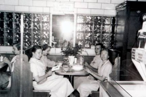 A black-and-white photo of four people sitting at a table in a diner, enjoying food and drinks.