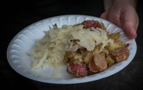 A hand holding a paper plate with sauerkraut and a mix of sausage and potatoes.