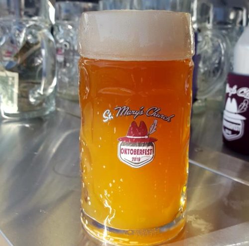 A frothy beer in a glass stein with an Oktoberfest logo, set against a blurred background of beer mugs.