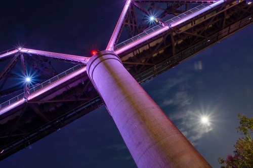A brightly lit bridge at night, with a full moon visible in the sky and colorful lights illuminating its structure.