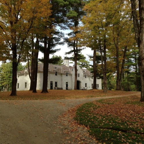 A scenic view of a white house surrounded by tall trees with autumn leaves on the ground.