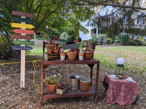 A wooden table with potted plants, a watering can, and a crystal ball, surrounded by signs pointing to various locations.