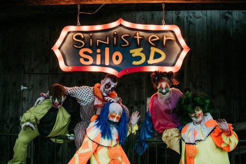 Five clowns in colorful costumes pose under a glowing sign that reads "Sinister Silo 3D" in a dimly lit setting.