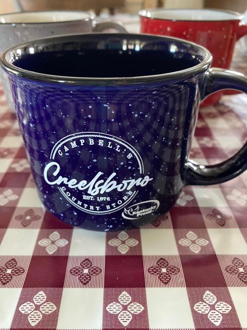 A blue mug with a white logo reading "Campbell's Creekshop Country Store" on a checkered tablecloth.