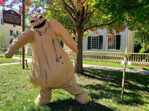 A large, smiling scarecrow stands in a grassy area with colorful leaves and a house in the background.