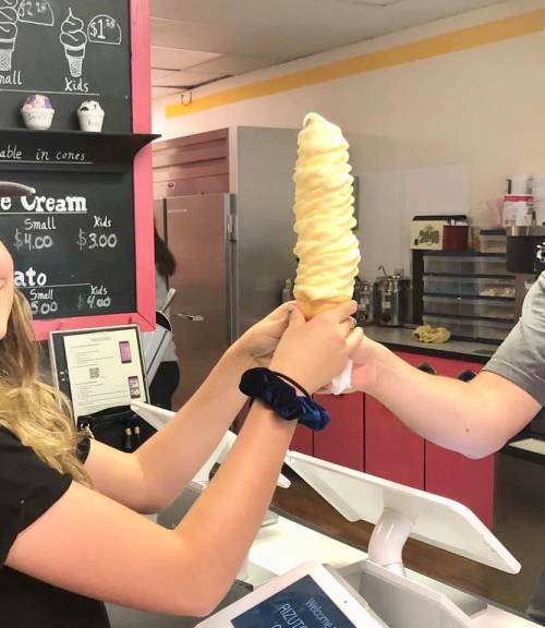 A person holds a large soft serve ice cream cone at a shop, with another hand reaching out to take it.