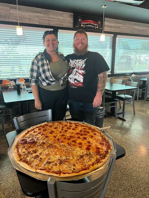 A man and woman stand beside a large pizza with pepperoni and sausage in a restaurant setting.
