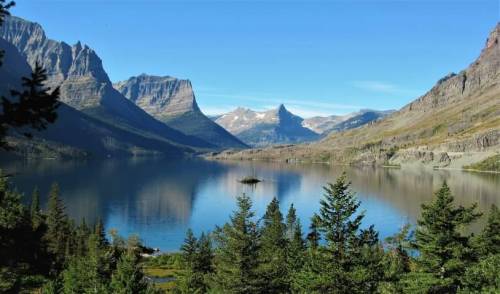 A serene lake surrounded by mountains, reflecting the landscape and framed by lush green trees.