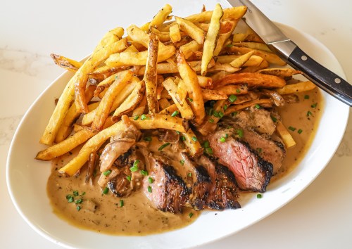 A plate of sliced steak in creamy sauce, served with a generous portion of golden French fries and garnished with chives.