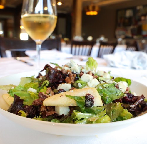 A fresh salad with mixed greens, pears, blue cheese, and nuts, served with a glass of white wine in a restaurant setting.