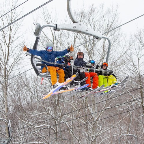 A group of four skiers on a chairlift, smiling and enjoying the snowy mountain scenery around them.